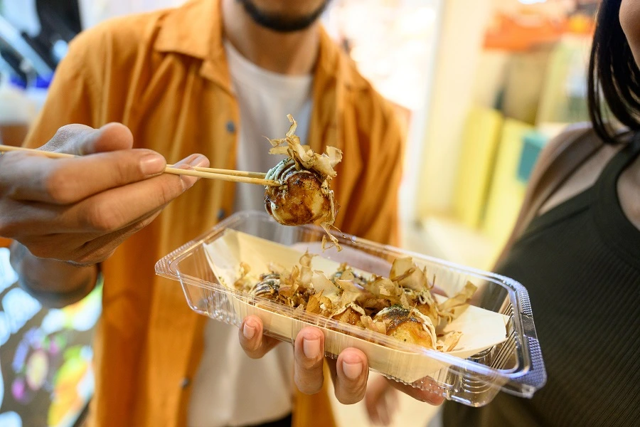 man in yellow shirt holding out takoyaki with chopsticks